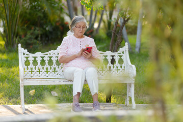 An older gray-haired woman with glasses is seated on a park bench, engrossed in her smartphone