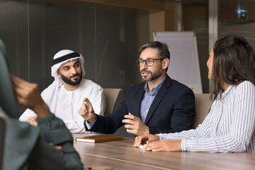 Professional meeting of four multicultural partners engaged in formal discussion in boardroom. European businessman consulting Arabian shareholders, share business strategy, explain project details