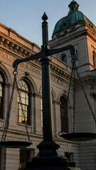 A black scale representing justice stands prominently in front of a grand courthouse with classical design elements. The structure features large windows and decorative details.