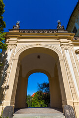 A low-angle shot of an arched gateway with decorative statues atop, framing a view of trees and...