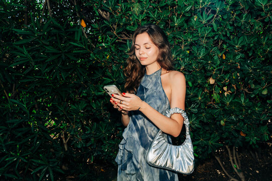 A fashionable woman checks her smartphone while smiling, standing against lush green bushes, dressed in a trendy summer outfit