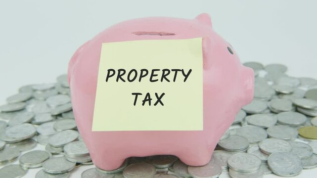 Property Tax text on yellow note paper with a hand inserting coins into a pink piggy bank, surrounded by coins on a white background