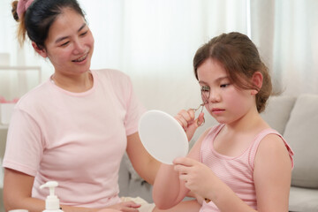 Caucasian girl learning makeup with Asian mom. Little girl using eyelash curler while looking into mirror. Mother smiling warmly, watching in cozy home setting with beauty products on table.