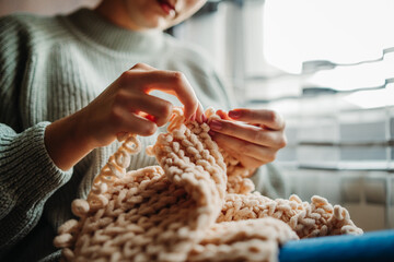 Young caucasian woman knitting blanket with her fingers by the window	
