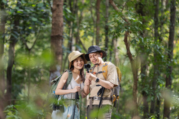 A couple of adventurous hikers explore the lush green forest while documenting their journey with a handheld camera. They wear outdoor gear, backpacks, and trekking poles, capturing memorable moments.