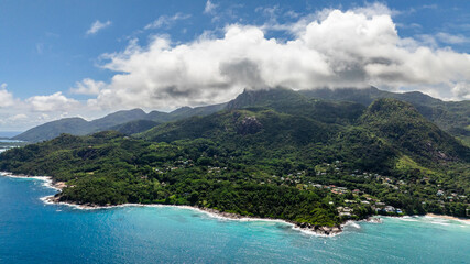 Green hills with clouds touching the peaks overlook turquoise waters along the coastline. Seychelles, Mahe.