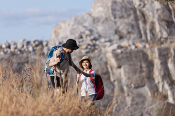A smiling woman helps her partner climb up a grassy hill during a hike, set against a rocky...