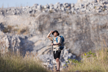 A male hiker with a backpack and trekking pole pauses on a rugged mountain trail, shielding his eyes from the sun. 