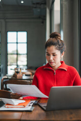 Woman in a red sweater working on a laptop in a modern cafe, reviewing documents with focused...