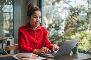 Focused woman in a red sweater using a laptop at a cafe table, surrounded by greenery and natural light.
