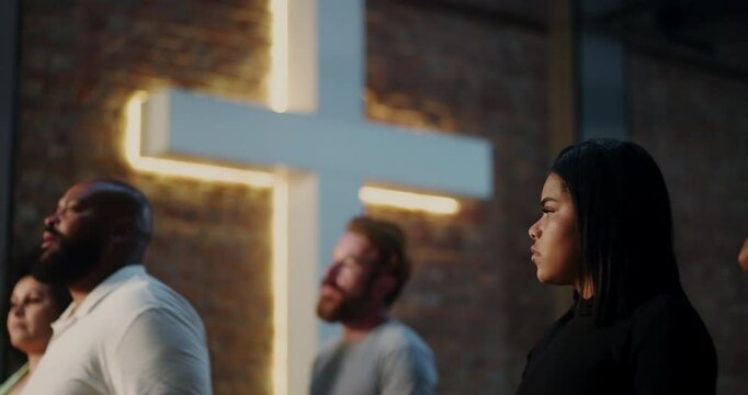 Young woman in black with serious expression, illuminated cross in background, spiritual gathering emphasizing Christian faith, contemplation, and diverse congregation