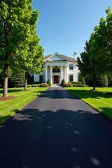 Asphalt driveway leading to a stately white home with classic columns, lush green lawn, and mature trees framing the entrance.