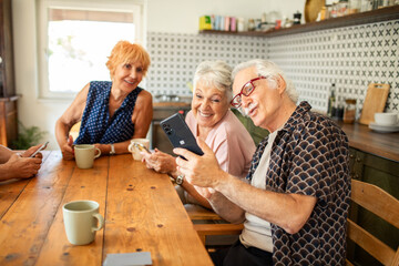 Happy senior friends using smartphone and drinking coffee in cozy kitchen