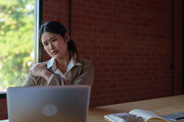 A businesswoman in a beige blazer sits at her desk, holding her shoulder with a pained expression,...