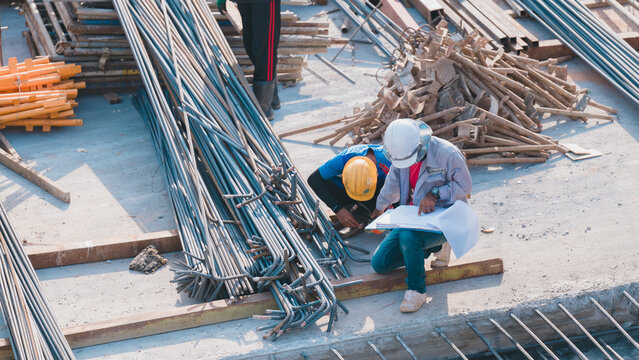 Construction Workers Collaborating on Site, Two construction workers review blueprints amidst piles of materials on construction site, ensuring accurate project planning and execution