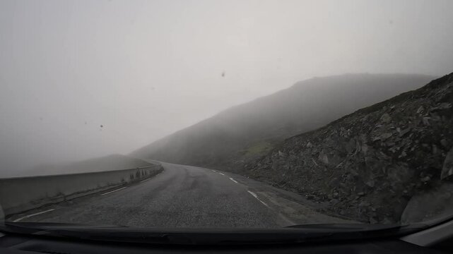 Driving on the E69 road through the tundra in northern Norway. Misty hills covered with grass and scattered stones surround the route, creating a remote and atmospheric landscape.