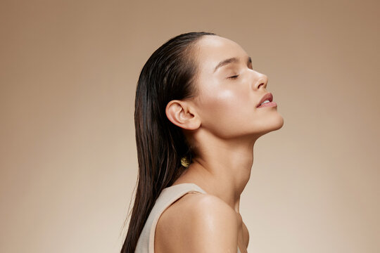 A serene young woman with long, wet hair poses against a neutral background, embodying beauty and tranquility through her peaceful expression and natural skincare routine