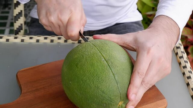 Male hands cutting pomelo fruit peel by kitchen knife on table close up.