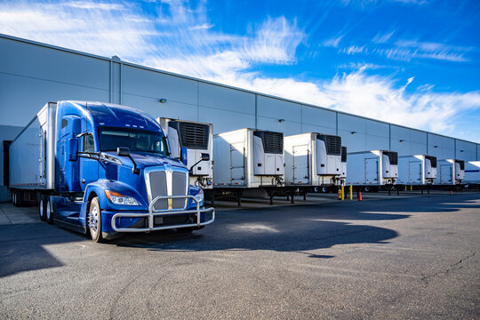 Big rig blue semi truck with refrigerator semi trailer standing in row with another semi trailers in warehouse dock gates loading cargo for the next freight