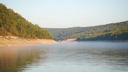 Obraz premium Misty lake surrounded by green forested hills at early morning calm water with light fog
