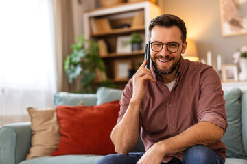 Young man is sitting on a sofa at home, engaged in a phone conversation. The scene captures a...