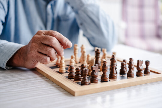Close up on chess board and wrinkled hands of senior man playing alone with chess on wooden table. Pastime in retirement lifestyle, strategy and problem solving - Powered by Adobe