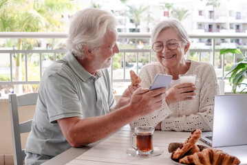 Smiling bonding senior retired couple shares news on mobile phone while enjoy breakfast together sitting outdoors on home terrace. E-commerce, social, tech concept