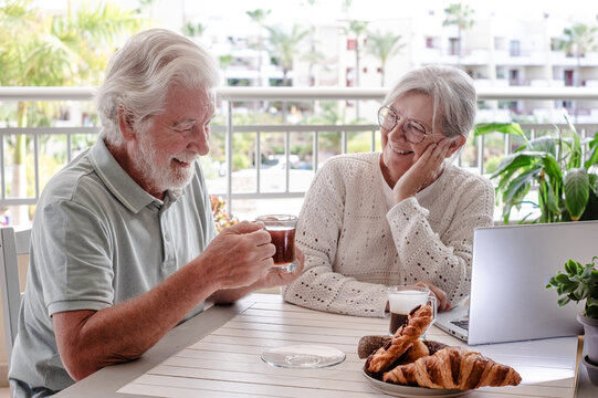Smiling bonding senior retired couple browsing with laptop while enjoy breakfast together sitting outdoors on home terrace. E-commerce, social, tech concept