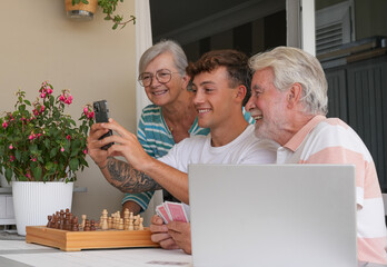 Happy multi generation family enjoying free time together sitting outdoors on terrace smiling playing cards and chess. Grandparents and young grandson stay together in leisure activity