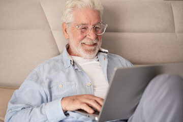 Fototapeta premium Smiling handsome caucasian senior bearded man with eyeglasses lying on bed using computer, elderly retiree browsing internet by laptop