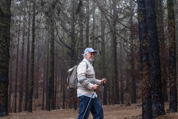 Leisure activity for an old senior man holding a backpack walking in mountain forest in a foggy winter day with the help of poles enjoying nature, freedom and free time. Bare trees in background