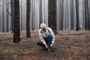 Fototapeta premium Active happy senior retired man with backpack walking in mountain forest on a foggy day stops to tie his shoes. Bare trees on backgrounds