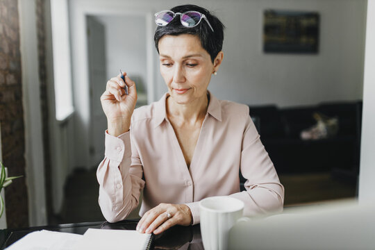 Female teacher of 50s checking homework of her student in copybook, correcting mistakes, holding pen in hand. Woman school principal making notes or looking through plan list in her notebook at table