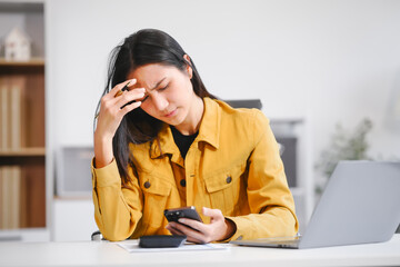 A businesswoman works at her desk, looking at her phone and documents. She feels stressed and...