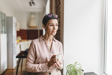 Concerned pensive worried female of 50s in elegant clothes with glasses on head holding cup of hot...