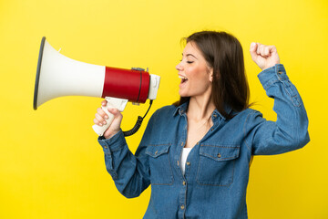 Young caucasian woman isolated on yellow background shouting through a megaphone to announce something in lateral position