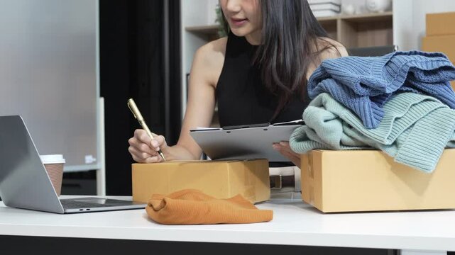 An Asian female business owner records a live vlog, showing a parcel box in front of a smartphone. She sells clothes online, engaging customers and promoting products for successful e-commerce sales