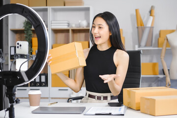 An Asian female business owner records a live vlog, showing a parcel box in front of a smartphone. She sells clothes online, engaging customers and promoting products for successful e-commerce sales