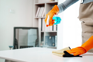 A young woman, wearing an apron, holds a spray bottle and microfiber cloth, preparing to clean a computer screen and check her desk at work, ensuring a clean and organized workspace