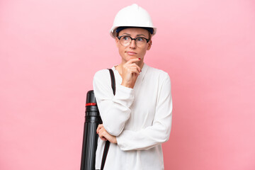 Young architect Russian woman with helmet and holding blueprints isolated on pink background having doubts and thinking