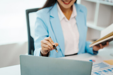 An Asian female accountant works happily at her desk in the office, using a laptop to analyze financial charts. She manages business finance and online transactions for company success