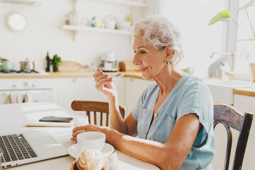 Side view of adorable caucasian female pensioner enjoying leisure time, breakfast and her favorite series sitting in front of laptop at kitchen table, looking at screen with smile, eating yogurt