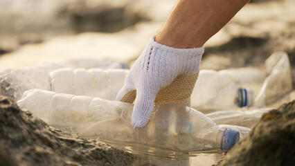 Gloved hand picking up a plastic bottle on the ocean coastline for a coastal cleanup in a recycling...