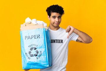 Young Venezuelan man holding a recycling bag full of paper to recycle showing thumb down with...
