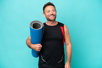 Middle age sport man going to yoga classes while holding a mat isolated on blue background thinking...