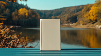 A blank white book cover stands upright on a wooden table by a tranquil lake. The backdrop features colorful autumn trees, creating a peaceful atmosphere perfect for creative presentations