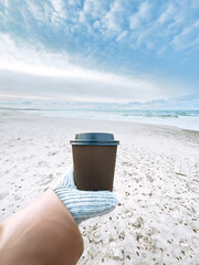 Female hand holding a brown mug with a hot drink, with a backdrop of sea, beach, and sky, creating a cozy and relaxing coastal moment.