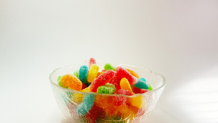 A vibrant assortment of sugar-coated gummy worms in various bright colors, including red, green, yellow, blue, and orange, placed in a transparent glass bowl. The candies have a soft, chewy texture