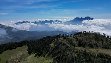 Aerial view natural beauty of green hills and meadow adorns the foreground. Mount Arjuno Welirang towers over thick white clouds. The sunlight casts a long shadow effect on the hillside.