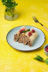 Thin crepes with poppy seed filling, decorated with raspberries and white chocolate, on a gray plate on a yellow concrete background.